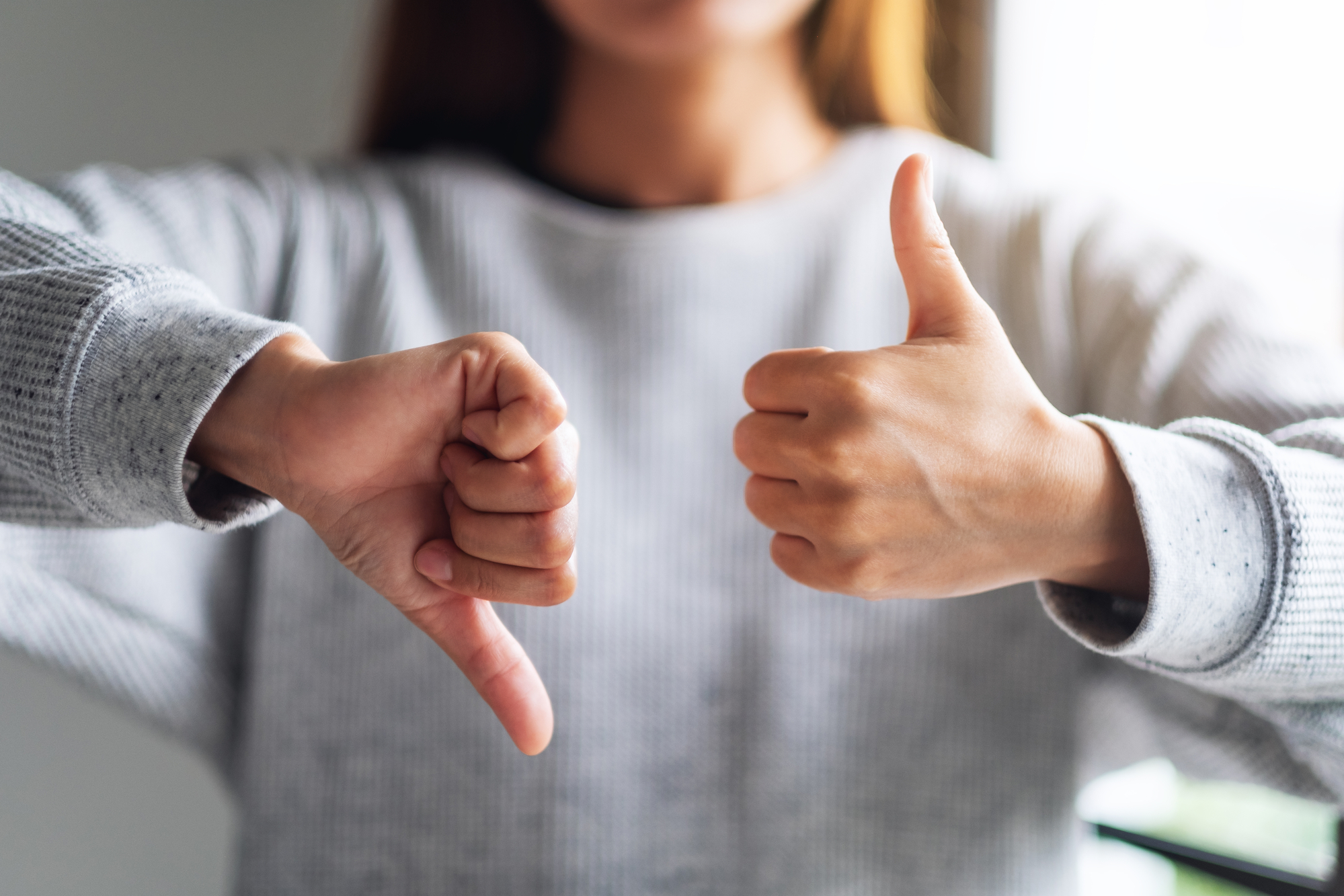 A person in a gray sweater gives a thumbs-up with their right hand while giving a thumbs-down with their left hand. The individual’s head and face are not visible in the image. The background is blurred.