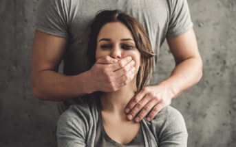 A seated woman looks forward with a man standing behind her, covering her mouth with his hand. He places his other hand on her shoulder. The woman wears a gray top, and the man is dressed in a light gray shirt. The background is a textured gray wall.