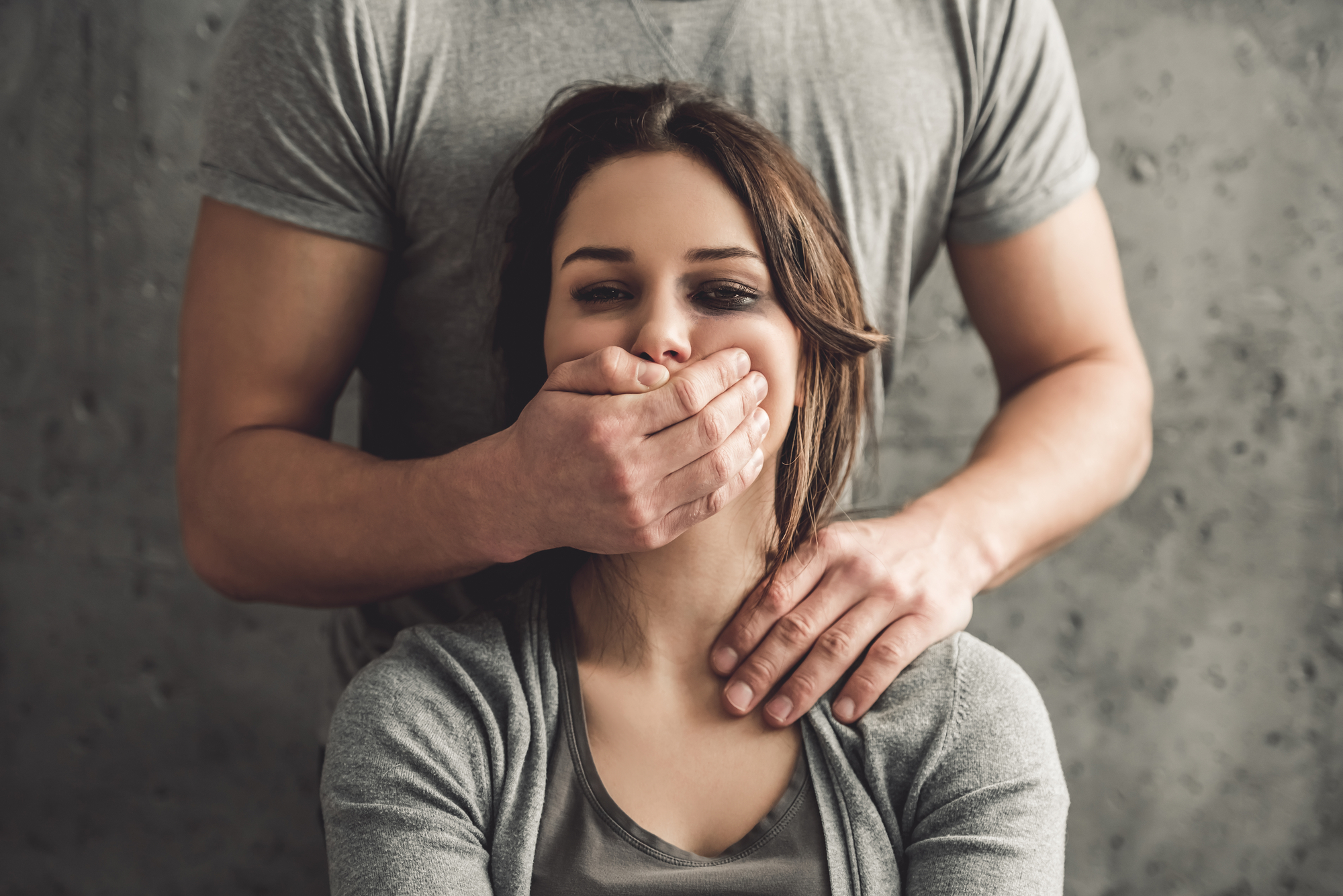 A seated woman looks forward with a man standing behind her, covering her mouth with his hand. He places his other hand on her shoulder. The woman wears a gray top, and the man is dressed in a light gray shirt. The background is a textured gray wall.