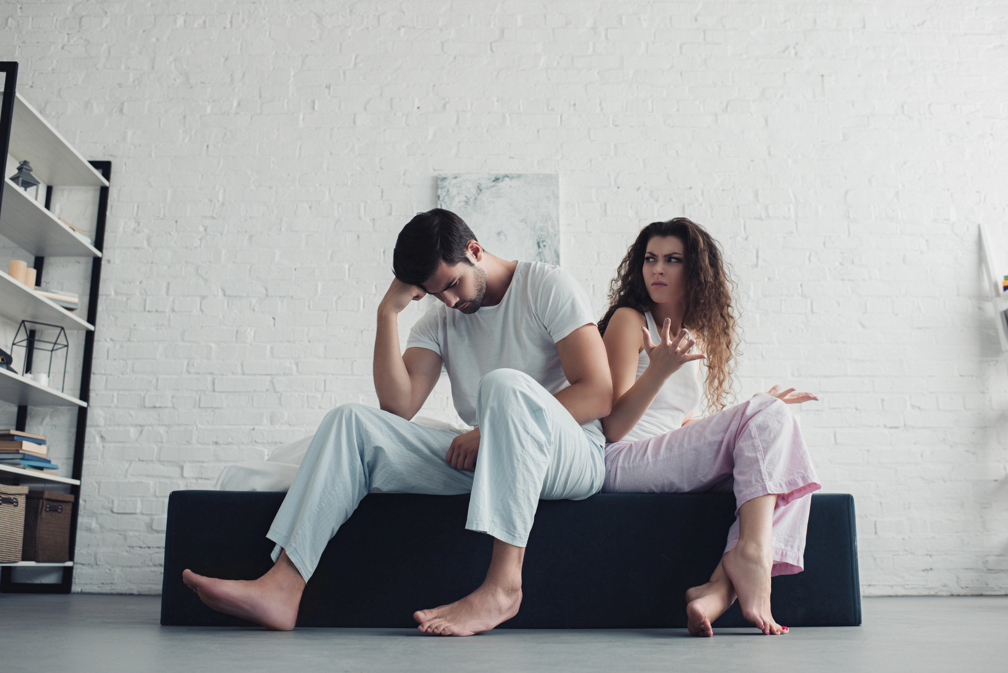 A man and a woman, both in pajamas, sit on a bed. The man looks down with his hand on his forehead, appearing distressed. The woman, sitting beside him, gestures with her hand, looking upset. The background features a white brick wall and minimalistic decor.