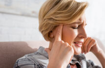 A middle-aged woman with short blonde hair sits on a couch, pressing her fingers to her temples and grimacing. She appears to be experiencing pain or discomfort, possibly a headache. She is wearing a plaid shirt. The background is blurred and bright.