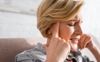 A middle-aged woman with short blonde hair sits on a couch, pressing her fingers to her temples and grimacing. She appears to be experiencing pain or discomfort, possibly a headache. She is wearing a plaid shirt. The background is blurred and bright.