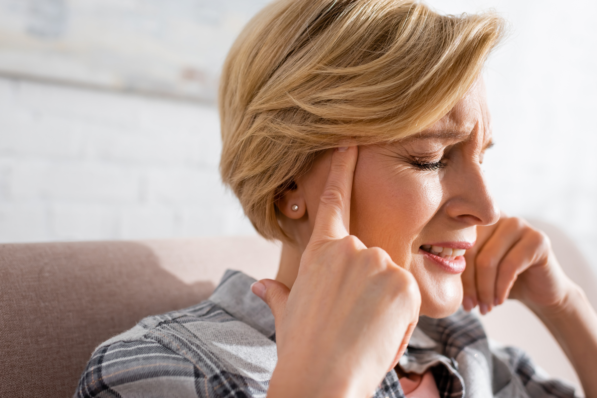 A middle-aged woman with short blonde hair sits on a couch, pressing her fingers to her temples and grimacing. She appears to be experiencing pain or discomfort, possibly a headache. She is wearing a plaid shirt. The background is blurred and bright.