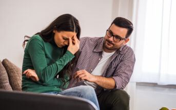 A woman sits on a couch, looking distressed with her hand on her forehead. Next to her, a man reaches out to comfort her with a concerned expression. They are in a bright room with large windows and a neutral-colored sofa.