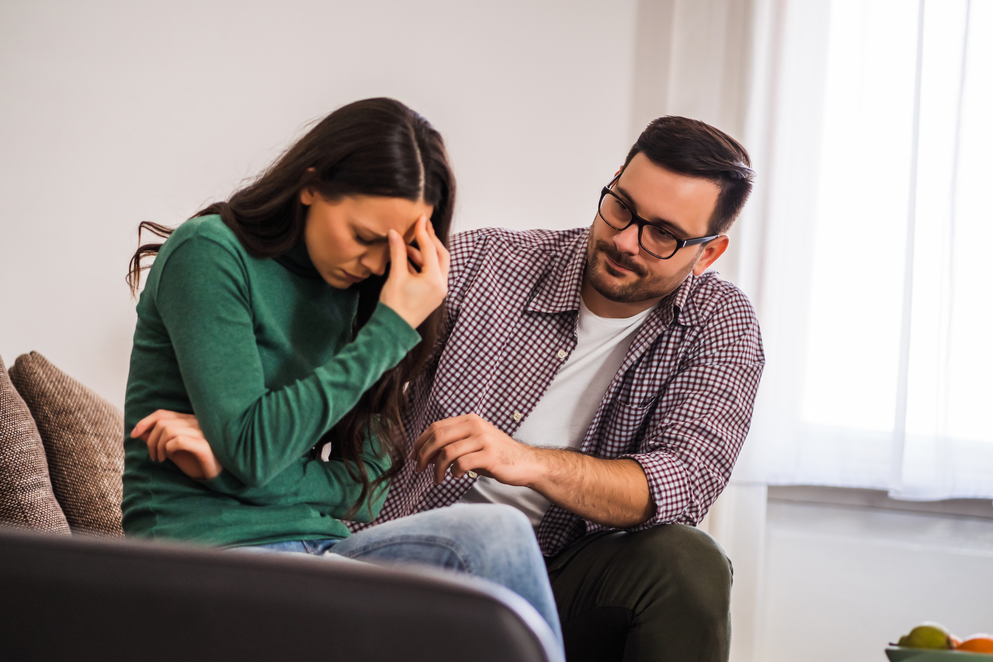 A woman sits on a couch, looking distressed with her hand on her forehead. Next to her, a man reaches out to comfort her with a concerned expression. They are in a bright room with large windows and a neutral-colored sofa.