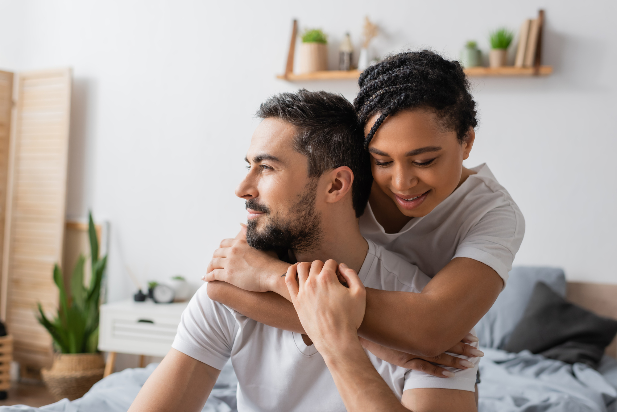 A couple in a cozy bedroom smiles and embraces. The woman hugs the man from behind as he looks to the side. Both wear white shirts. The room is decorated with plants, shelves, and neutral-colored furniture, creating a relaxed atmosphere.