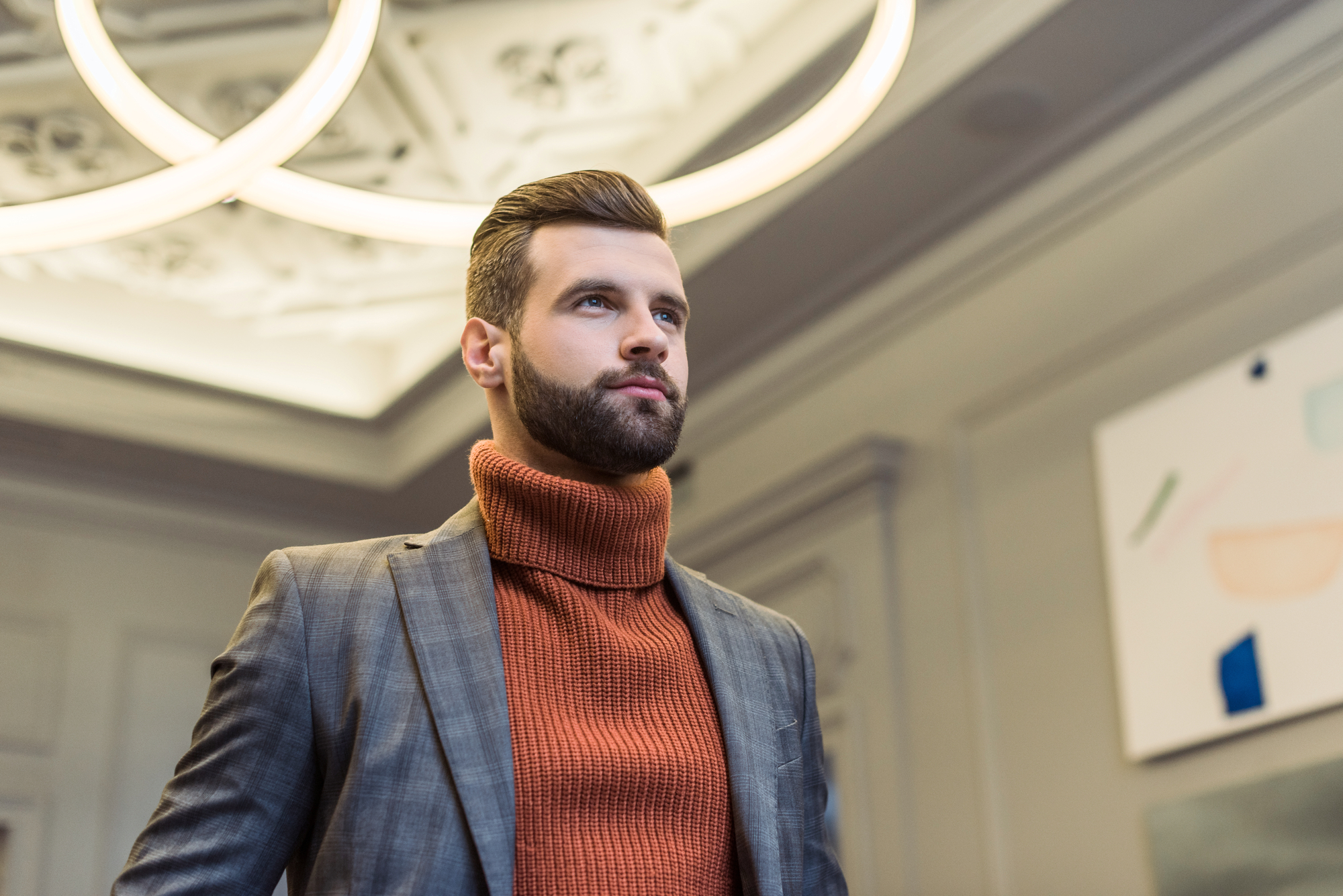 A bearded man with styled hair stands confidently, gazing off into the distance. He wears a gray checkered suit over a chunky rust-colored turtleneck sweater. The setting appears to be a modern room with artistic wall decor and circular ceiling lights.