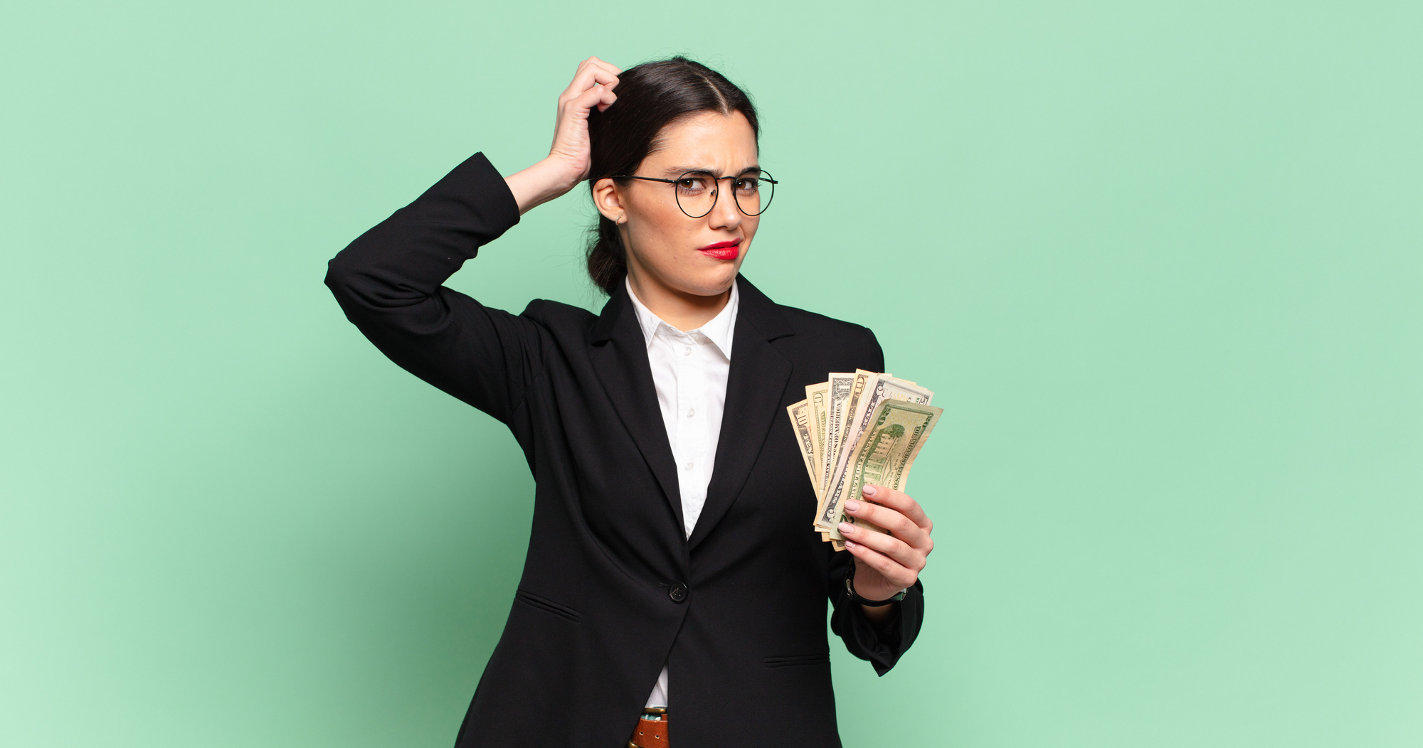 A person with long dark hair in a ponytail, wearing glasses, a white shirt, and a black blazer, holds several US dollar bills in one hand while scratching their head with the other. The background is a solid light green color. The person appears puzzled.