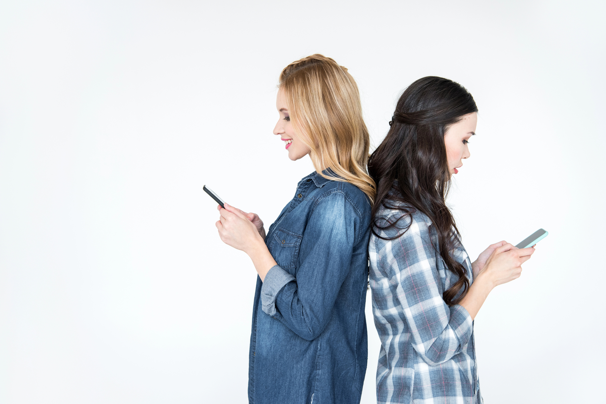 Two women standing back-to-back, both looking at their phones. The woman on the left has blonde hair and is wearing a denim shirt. The woman on the right has dark hair and is wearing a plaid shirt. They are against a plain white background.