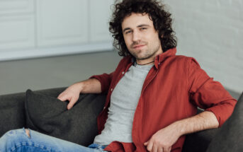 A young man with curly hair and a beard is sitting on a couch, leaning back with one arm resting on the backrest. He is wearing a red button-up shirt over a gray t-shirt and ripped blue jeans. He is looking at the camera with a relaxed expression.