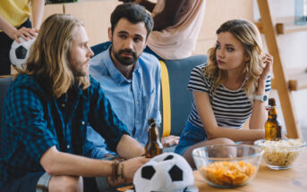 Three friends are sitting on a couch, engaged in conversation, with snacks and drinks on a table in front of them. Two soccer balls are placed on the couch. The group appears relaxed and focused, possibly watching a soccer game together.