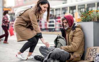 A woman in a tan coat kneels to give change to a homeless man sitting on the sidewalk. The man, wearing a red hoodie under a brown coat, has a sign beside him that reads "Once I was like you." A bus is visible in the background with people walking by.