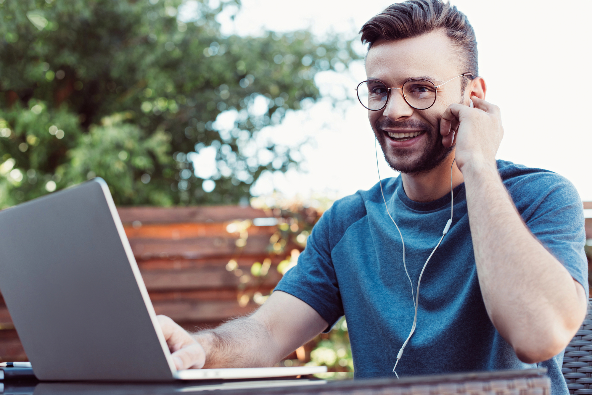 A young man with glasses, wearing a blue shirt, is smiling while working on a laptop outdoors. He has earphones in and is touching his ear with one hand. Background shows greenery and a wooden fence.