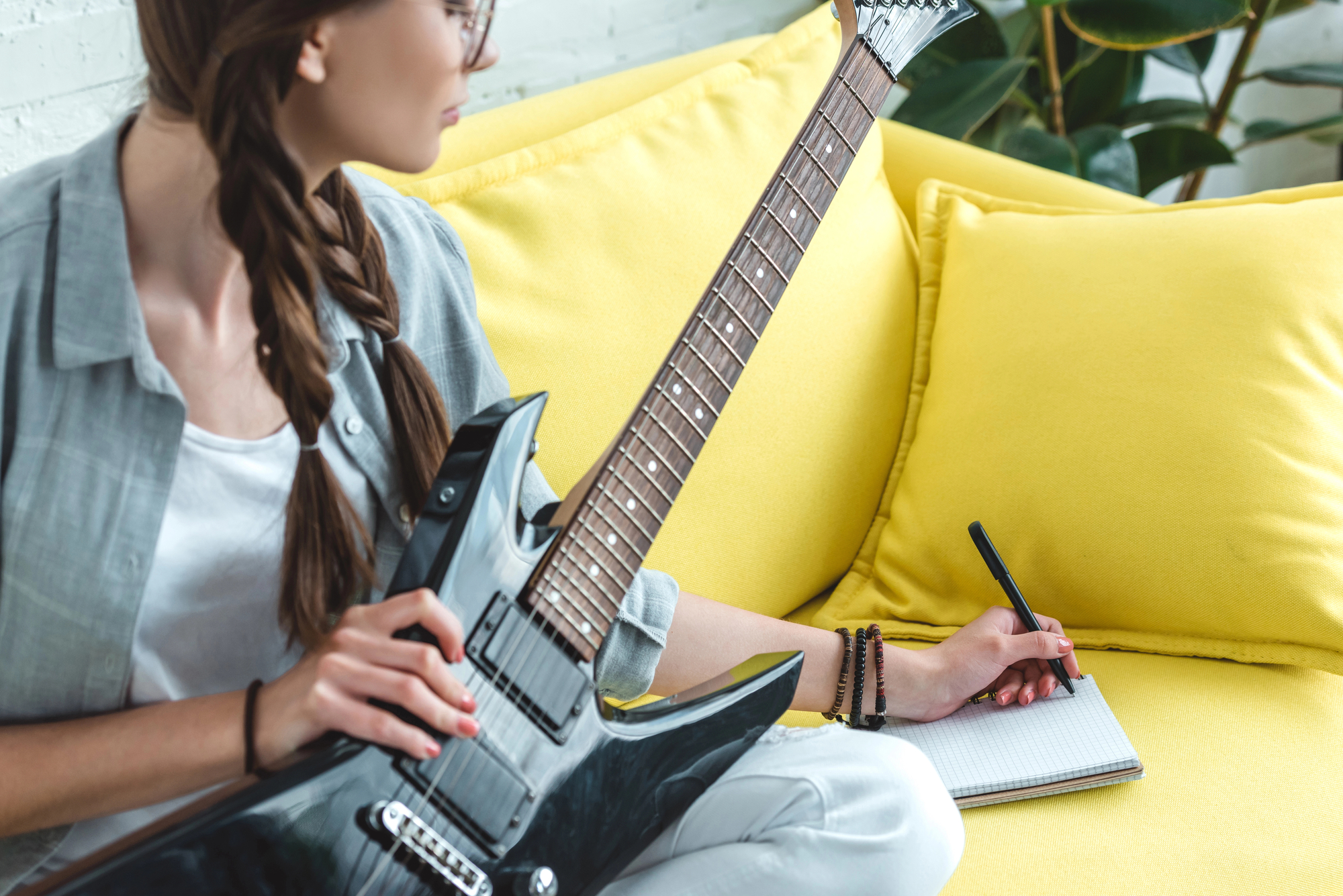 A person with a braid sits on a yellow couch, holding an electric guitar in one hand and writing on a notepad with a pen in the other hand. They are dressed casually, and a plant is visible in the background.