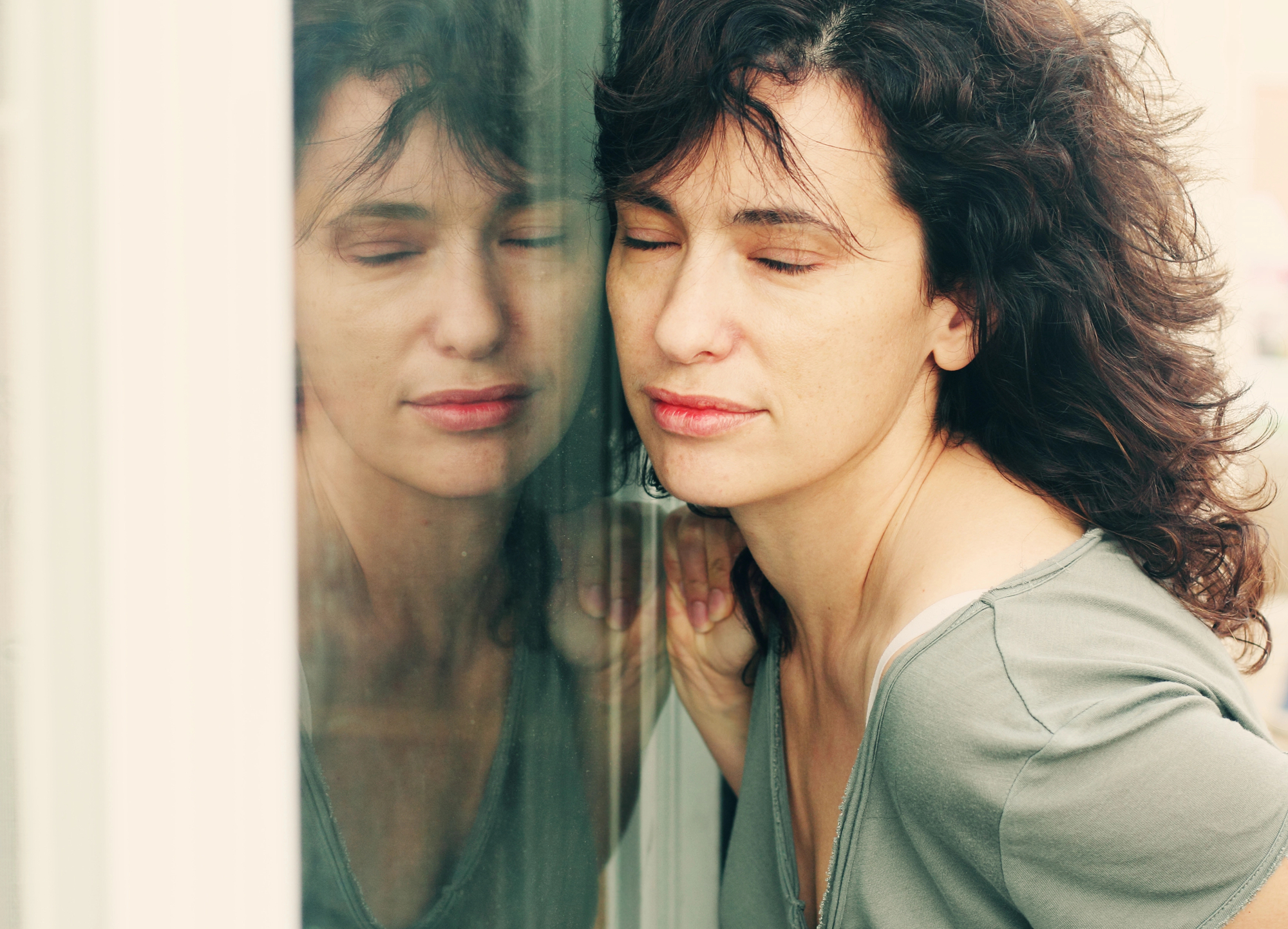 A woman with curly dark hair and a pale complexion rests her head against a window, eyes closed, with a calm expression. The reflection of her face is visible in the glass. She wears a light grey top, and the background is softly blurred.