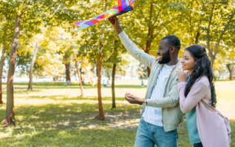 A man and woman are smiling as they get ready to fly a colorful kite in a sunlit park. The man holds the kite above his head while the woman stands close, looking at the kite. Trees with green foliage surround them, and soft sunlight filters through the branches.