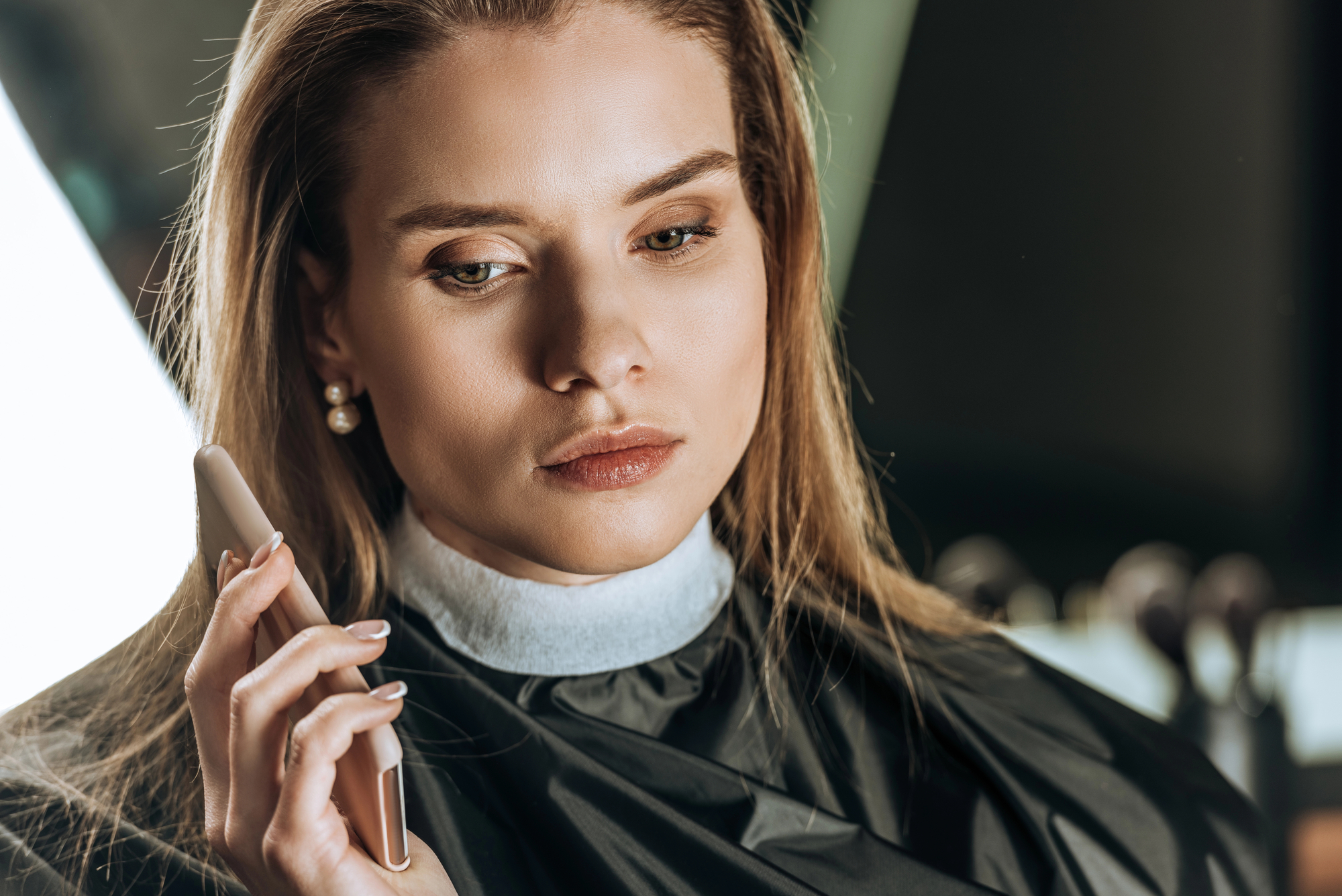 A woman with straight, blond hair wearing pearl earrings and a black salon cape holds a smartphone to her ear. She appears focused and thoughtful in a well-lit indoor setting.