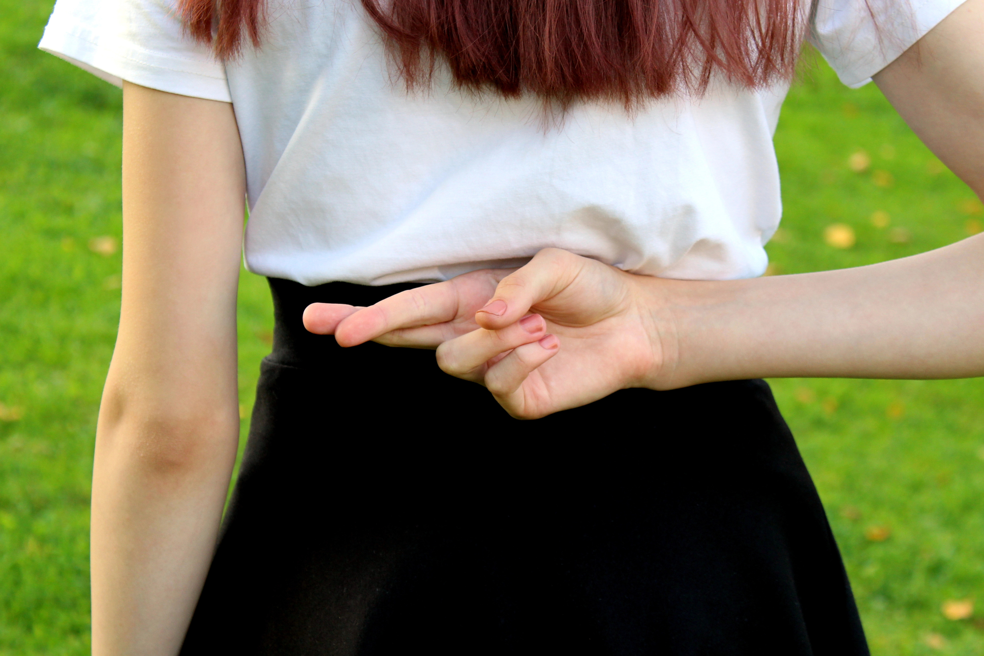 A person with shoulder-length brown hair is standing outside on green grass, wearing a white t-shirt and black skirt. They have their hand behind their back, fingers crossed.