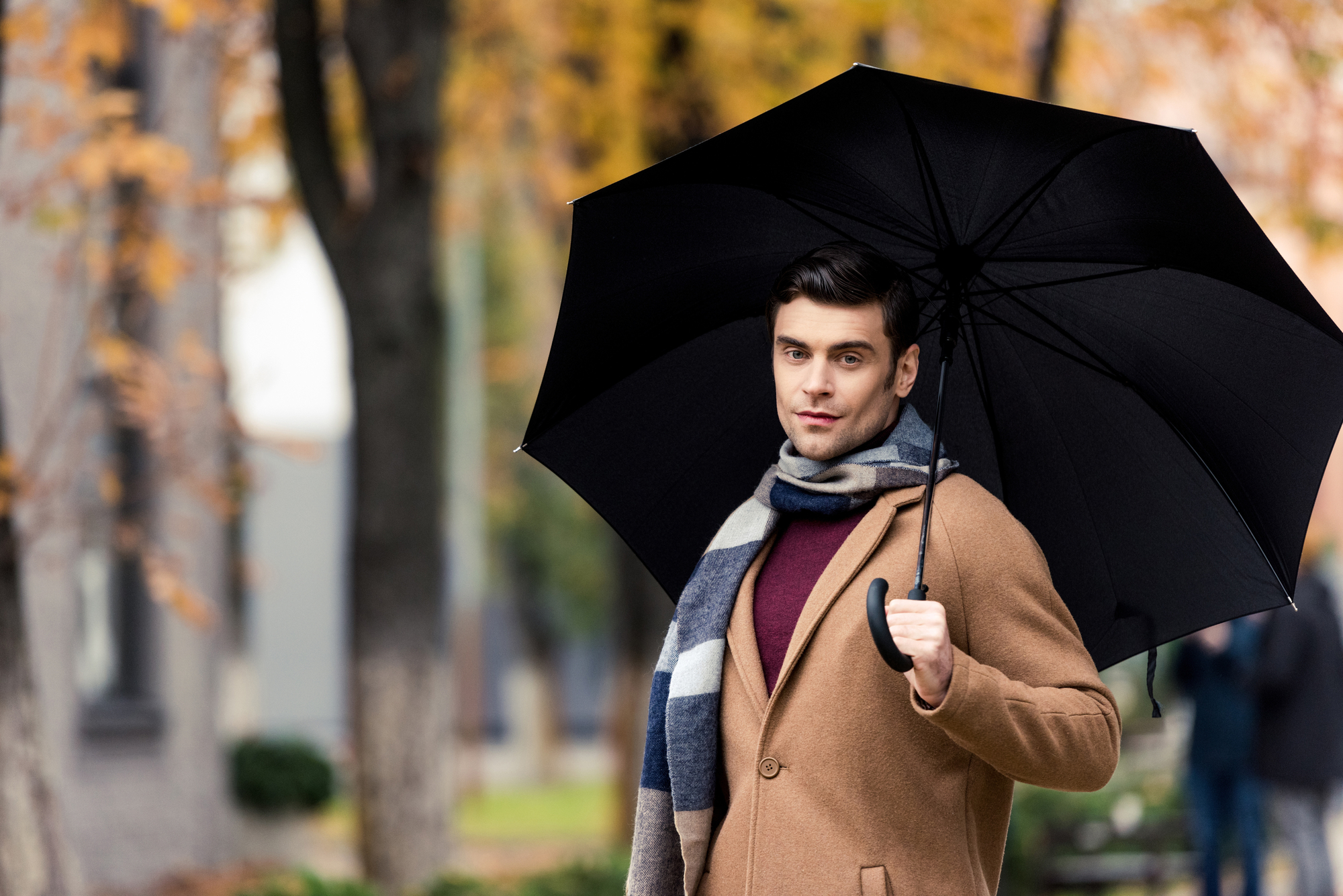 A man holding a black umbrella stands in a park during autumn. He's wearing a camel-colored coat, a scarf, and a burgundy sweater. The trees in the background have fall foliage, and the scene is slightly blurred.