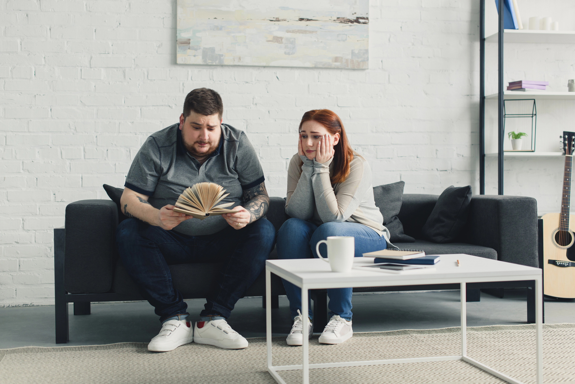 A man and woman sit on a sofa, looking concerned. The man holds an open book, while the woman rests her chin on her hands. The room features a coffee table with a white mug, a notebook, and a pen. A guitar leans against the wall, and a light-colored painting hangs above them.