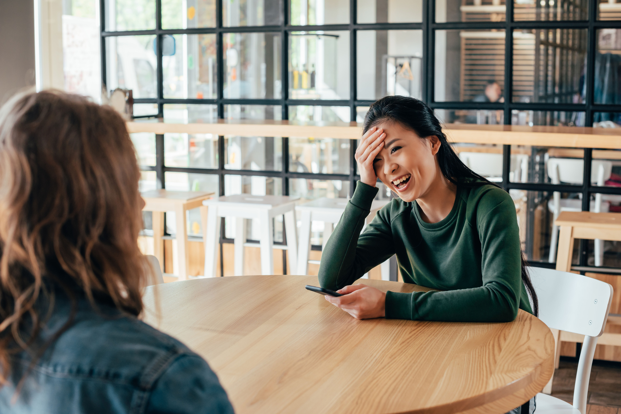 Two people are sitting at a round wooden table in a modern cafe with a glass wall in the background. One person, wearing a green top, is laughing with one hand on their forehead, while holding a phone in the other hand. The other person is facing away.