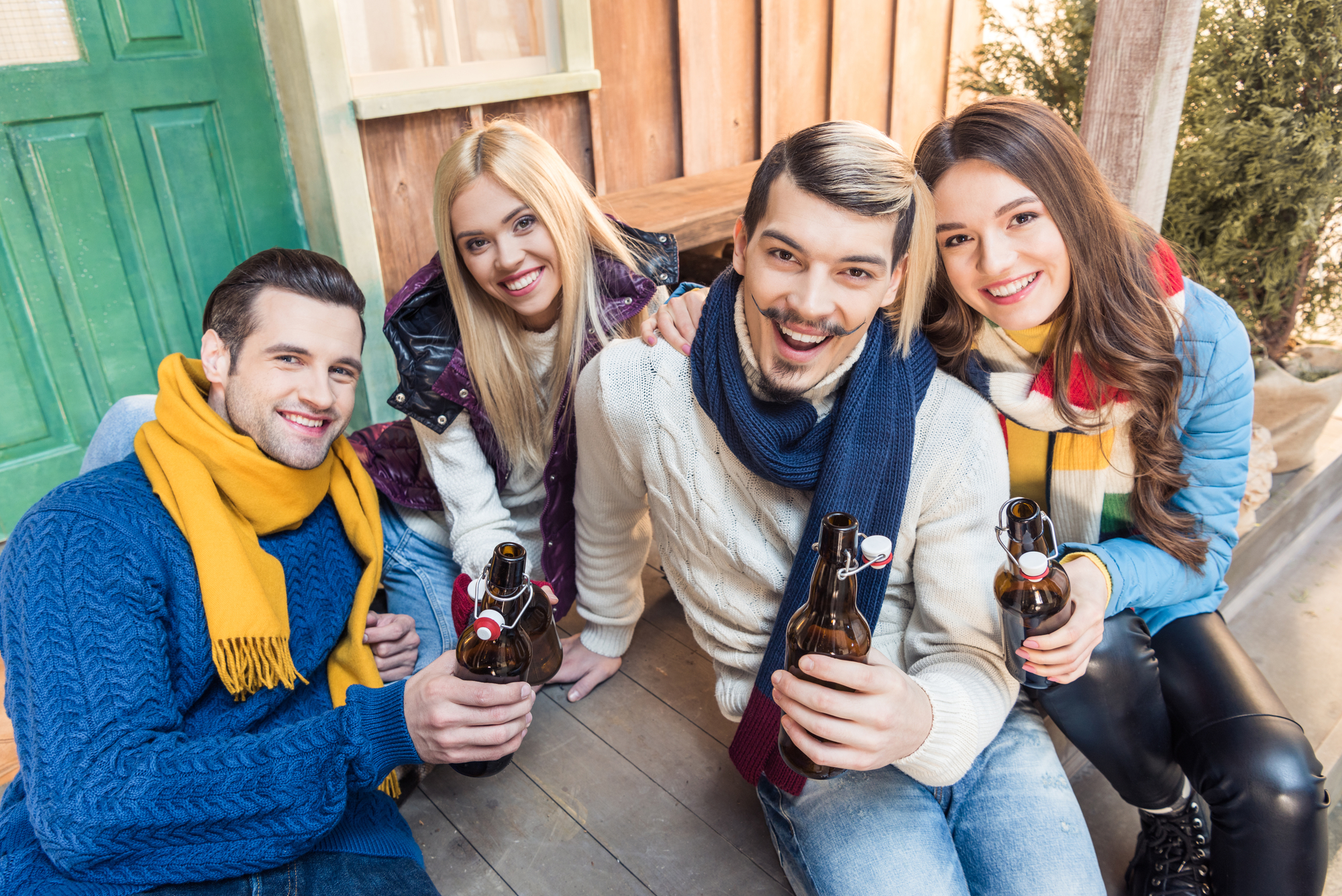 Four friends gathered outside, smiling and holding bottles of beer. They are warmly dressed in sweaters, scarves, and jackets, suggesting a chilly weather. The background shows a wooden cabin, adding a cozy, rustic feel to the scene.