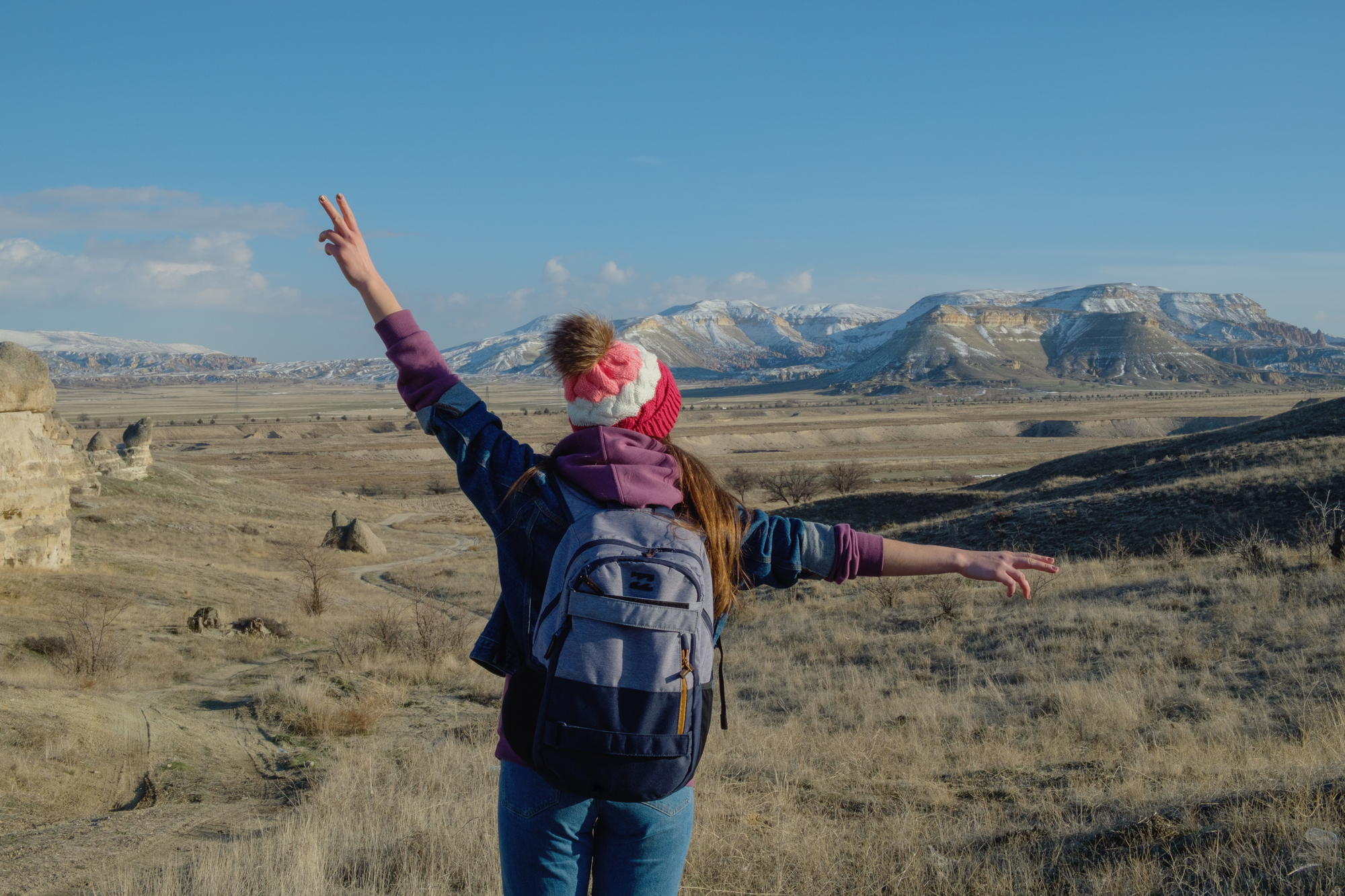 A person wearing a backpack and a red beanie stands in a wide-open, arid landscape with arms outstretched, giving a peace sign with one hand. Snow-capped mountains are visible in the background under a clear blue sky.