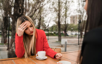 A woman with long blonde hair, wearing a red blazer, sits at an outdoor café table with a cup of coffee. She holds her head in her hand, appearing distressed. Another person, partially visible, sits across from her, also with a cup of coffee. Trees are in the background.