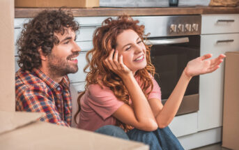 A man and woman sit on the floor of a kitchen with moving boxes around them. The man has curly hair and a beard, wearing a plaid shirt. The woman, with wavy hair, smiles and gestures with her hand, wearing a pink shirt. The setting suggests they are moving in together.