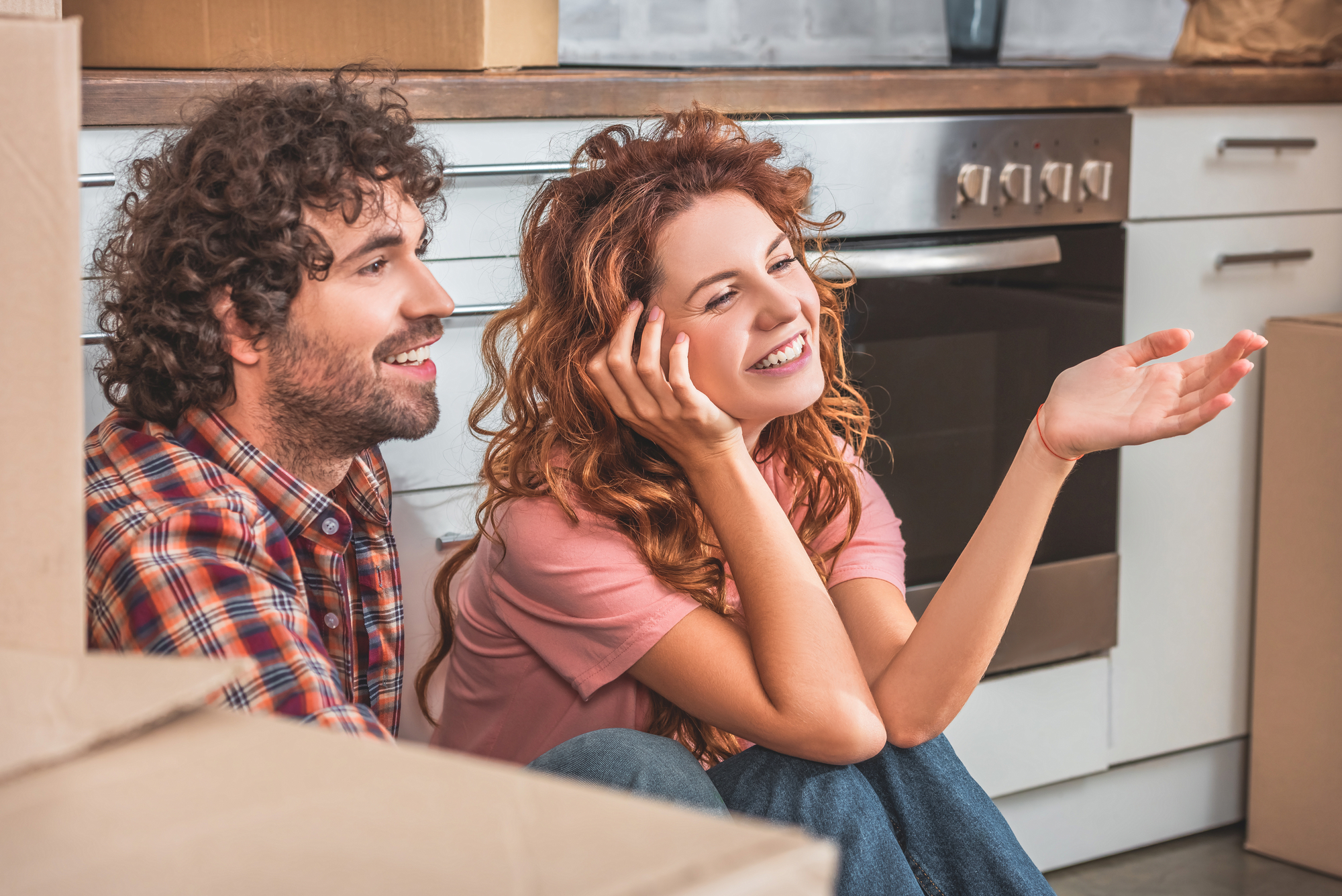 A man and woman sit on the floor of a kitchen with moving boxes around them. The man has curly hair and a beard, wearing a plaid shirt. The woman, with wavy hair, smiles and gestures with her hand, wearing a pink shirt. The setting suggests they are moving in together.