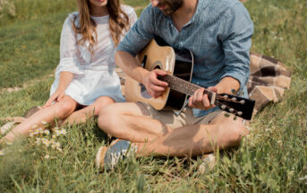 A man sitting on the grass playing an acoustic guitar, wearing a blue shirt and beige shorts. Next to him is a woman in a white dress, sitting on a blanket, smiling and listening. They are outdoors, surrounded by green grass and small wildflowers.