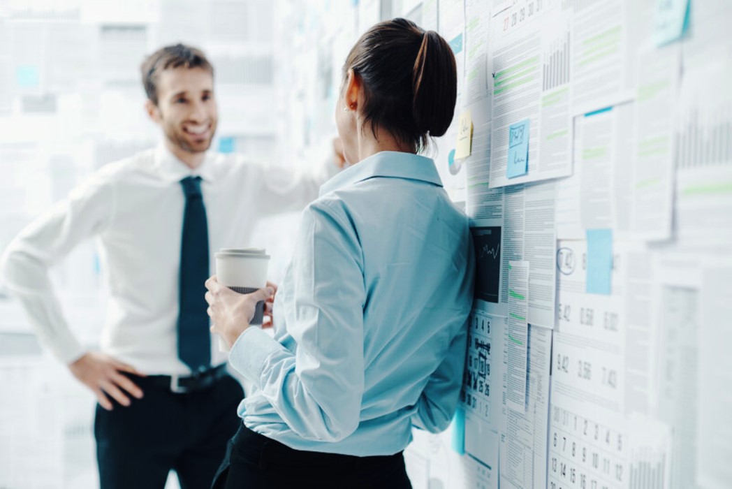 Two professionals, a man and a woman, engage in conversation in an office, standing in front of a wall covered with papers, charts, and post-it notes. The woman holds a coffee cup, while the man appears to be explaining something with a smile. Both are dressed in business attire.