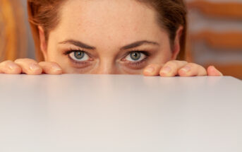 Close-up of a person with light-colored eyes peeking over the edge of a white surface. Only the top half of their face and hands gripping the surface are visible. The background is blurred, suggesting an indoor setting.