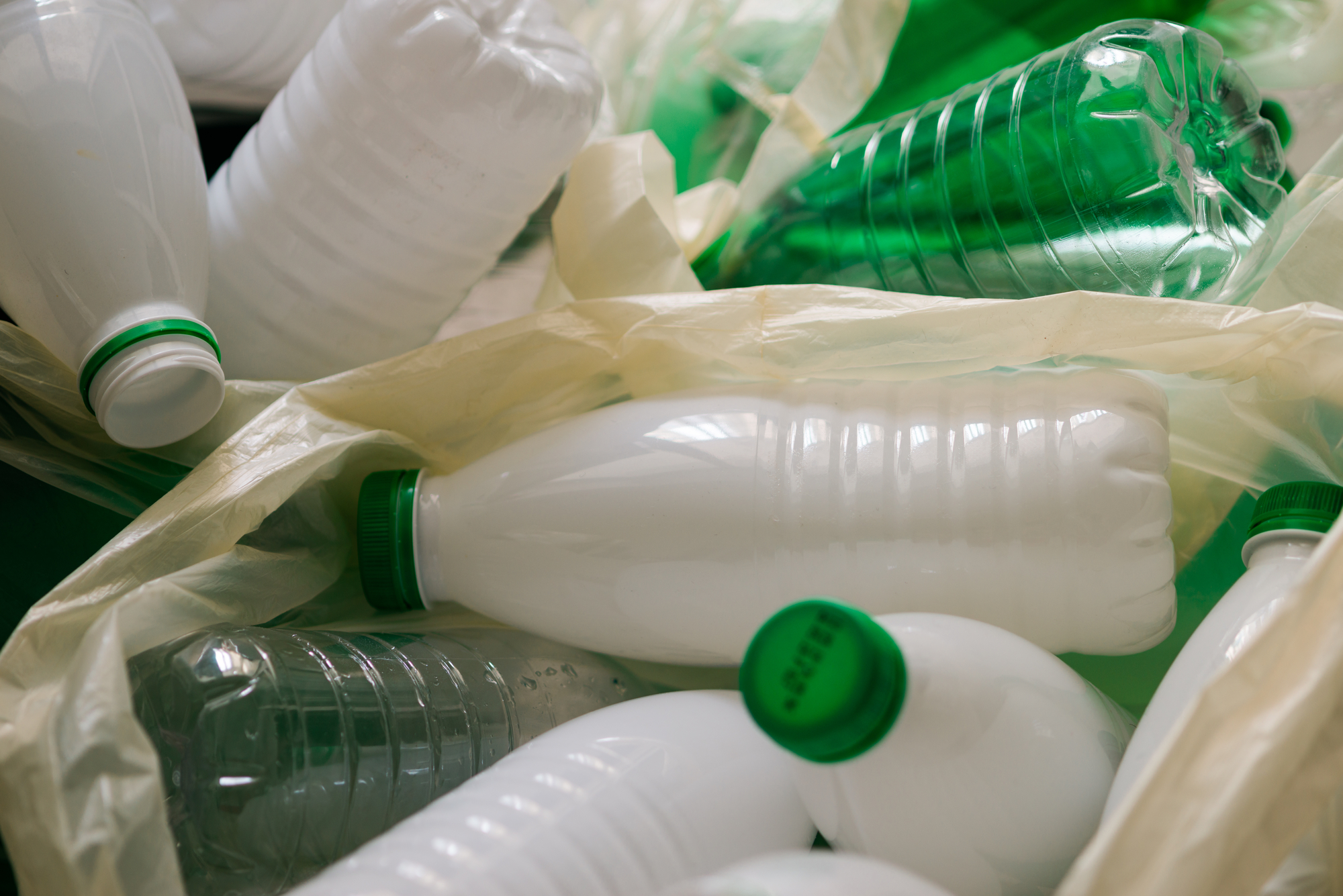 A collection of plastic bottles, including white and green ones, some partially wrapped in plastic bags. The bottles are arranged irregularly, some lying horizontally and others at different angles. The bottles appear to be empty.