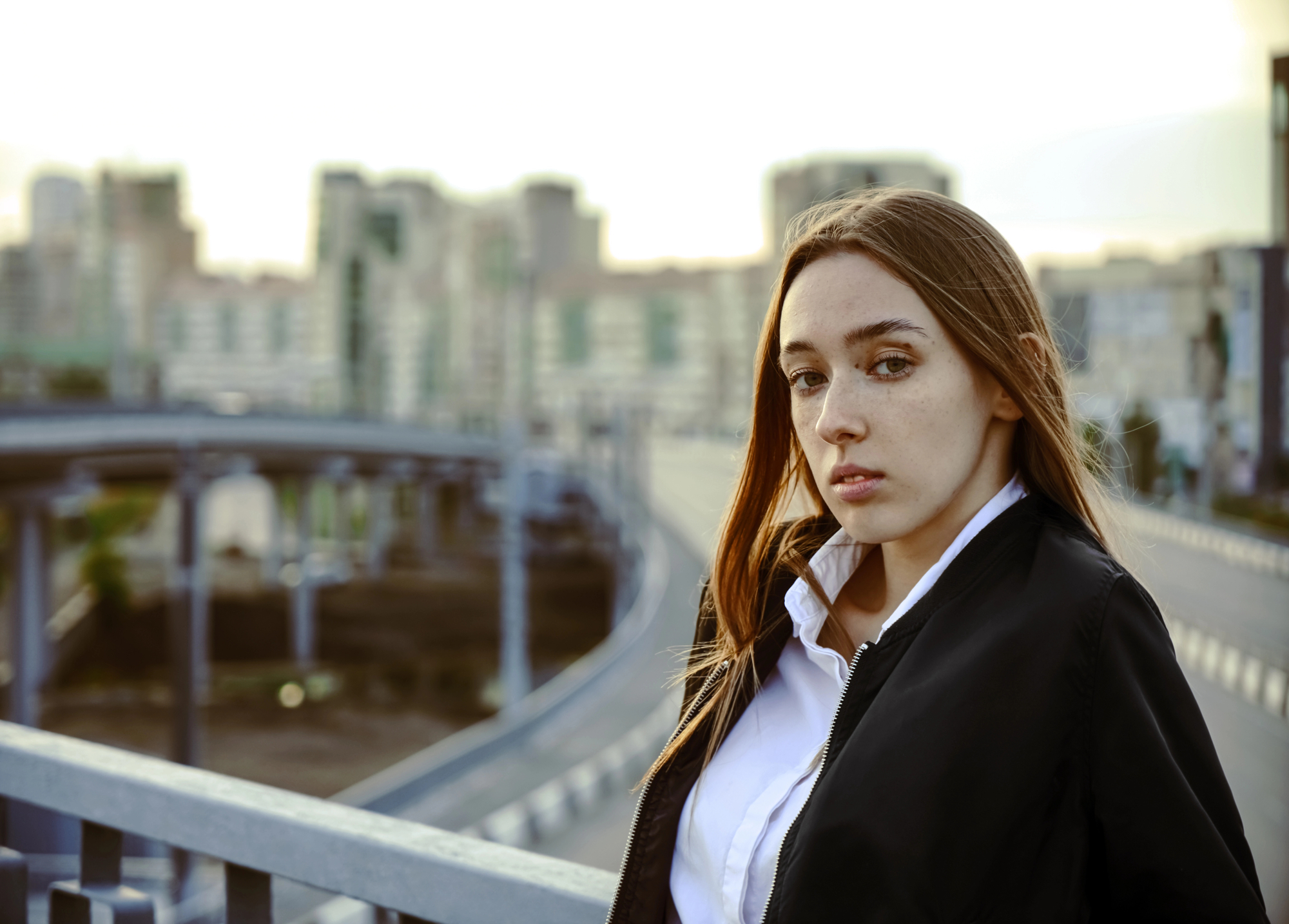 A young woman with long brown hair and serious expression stands on a bridge overlooking an urban cityscape with curved roads and tall buildings in the background. She is wearing a black jacket over a white shirt. The sky is overcast.