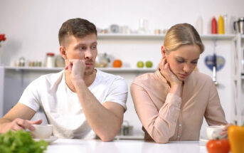 A man and woman sit at a table in a kitchen, both resting their chins on their hands with thoughtful, concerned expressions. The man looks at the woman, while she gazes downward. Various kitchen items are visible on shelves in the background.