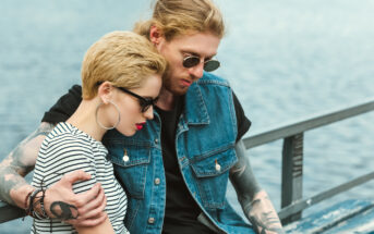 A couple sits close together on a bench by the water. The woman has short blond hair and wears glasses, hoop earrings, and a striped shirt. The man has long blond hair in a bun, wears sunglasses, and is dressed in a sleeveless denim jacket over a black shirt.