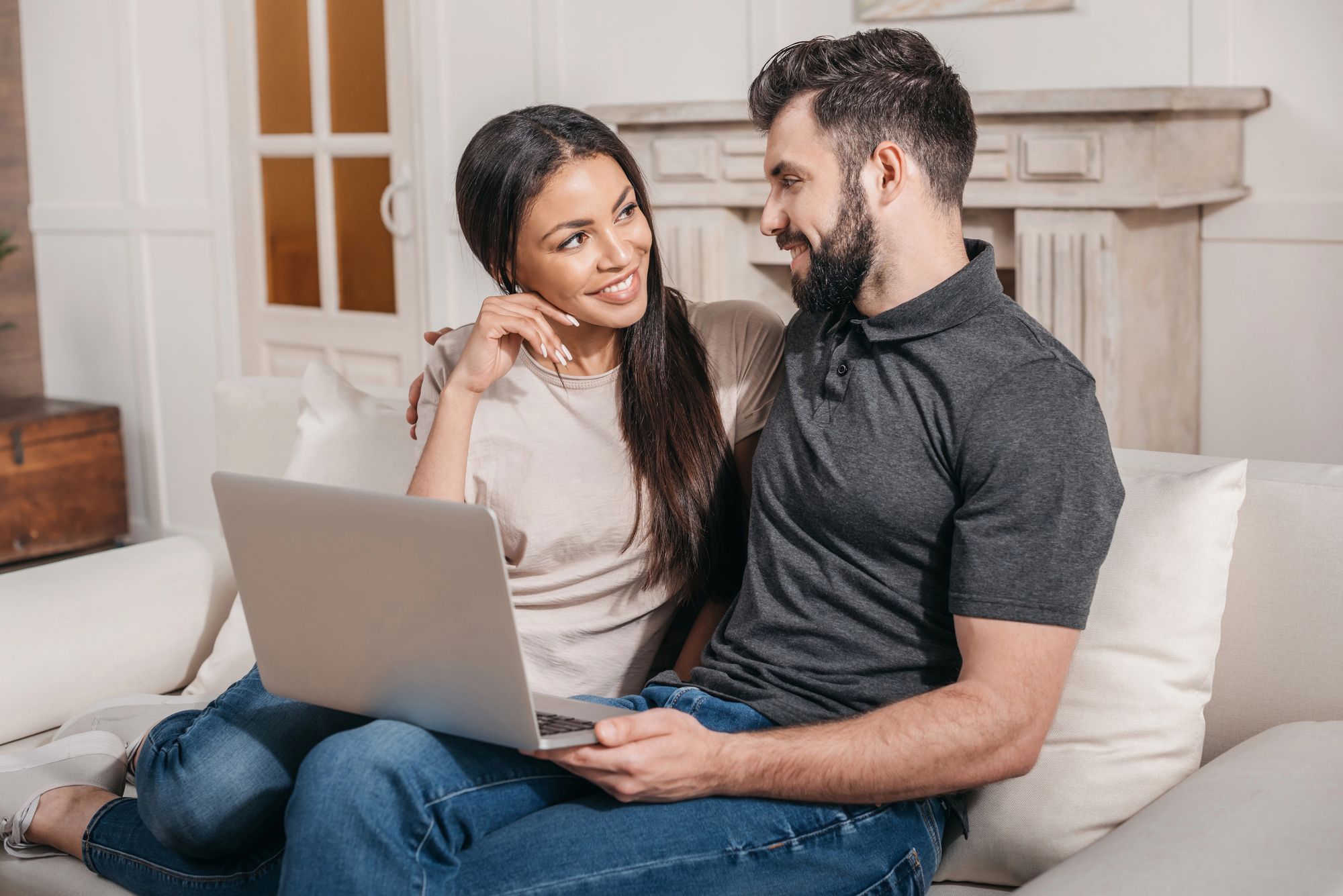 A smiling woman and man sit close together on a couch. The woman, with long dark hair, wears a beige shirt and blue jeans. The man, with short dark hair and a beard, wears a grey polo shirt and blue jeans, holding a laptop. They gaze at each other affectionately.