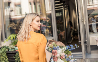 A woman with blonde hair, wearing a mustard yellow jacket, is smiling while holding a bouquet of colorful flowers. She is standing in front of an open storefront with plants and flowers visible within and outside the shop.