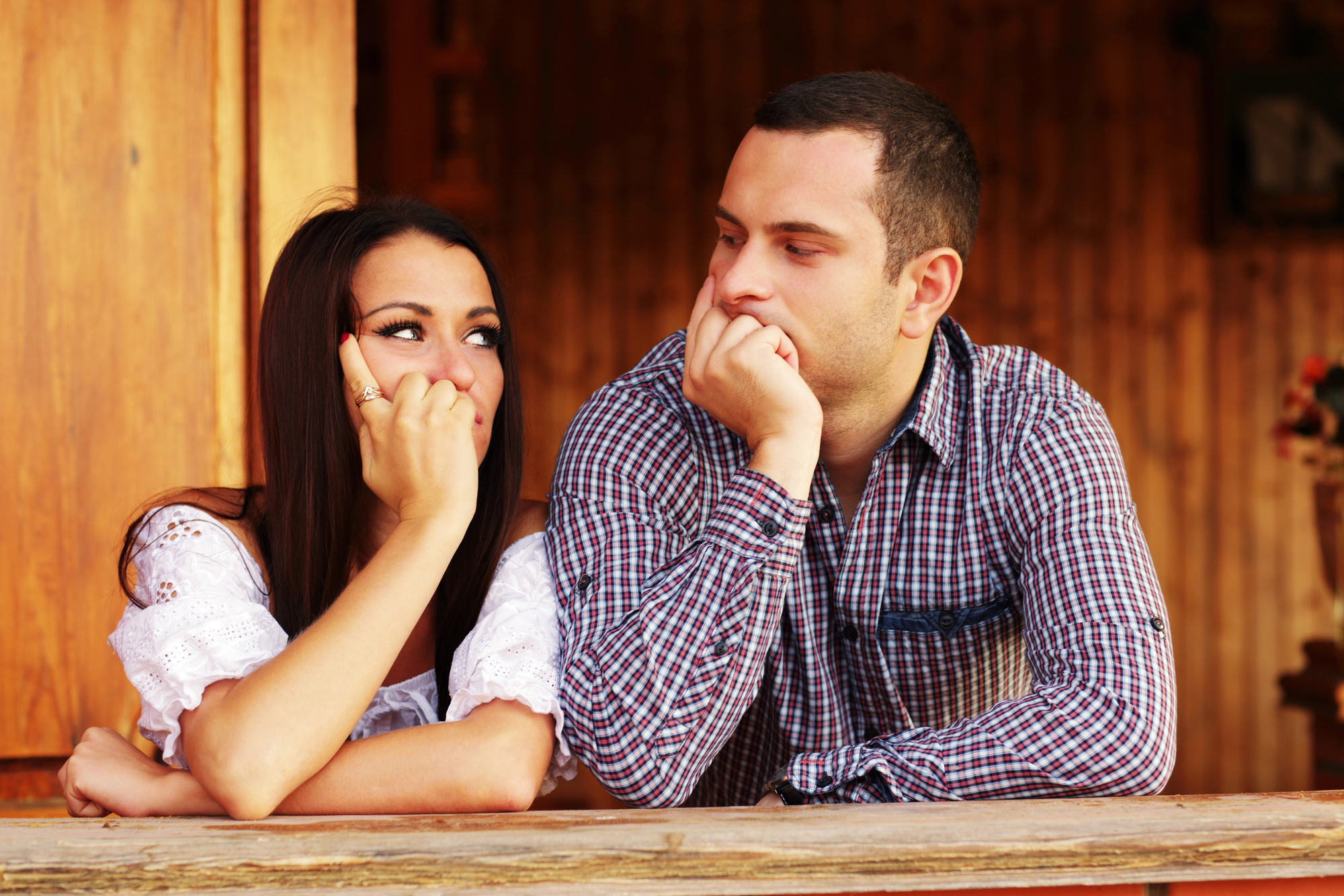 A woman and a man lean against a wooden railing, facing each other. The woman, with long dark hair and wearing a white top, looks thoughtful. The man, in a checkered shirt, rests his head on his hand and gazes at the woman. They appear to be in conversation.