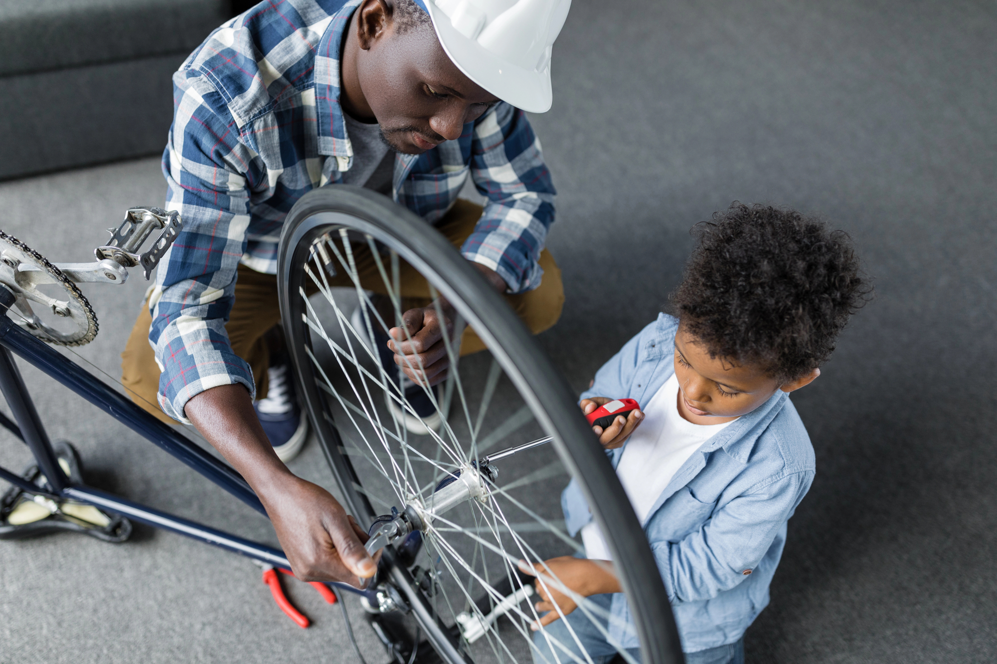 An adult wearing a hard hat assists a young child in repairing a bicycle wheel. The child, holding a red screwdriver, watches attentively while the adult adjusts the wheel components. Both are focused on the task and sit on a carpeted floor indoors.