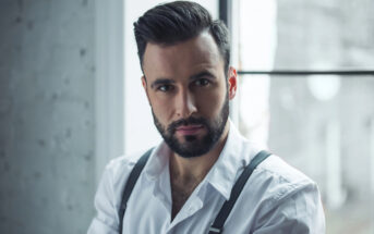 A man with a neatly trimmed beard and styled hair is wearing a white dress shirt with suspenders. He is posing in front of a window, which allows soft, natural light to illuminate his face and creates a serene ambiance in the room with a partially visible brick wall.