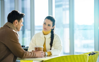 A man and woman sit at a table in a brightly lit cafe, engaged in conversation. The man, wearing a brown coat and scarf, holds a coffee cup. The woman, with a long braid and in a white jacket, listens attentively. Yellow chairs are visible in the foreground.