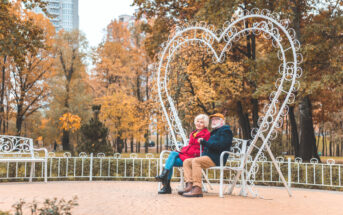 An elderly couple sits on a bench in a park with autumn foliage. They are under a large, heart-shaped metal frame. The woman, in a bright red coat, leans on the man, who is dressed in dark clothes and a cap. Both look content and peaceful.