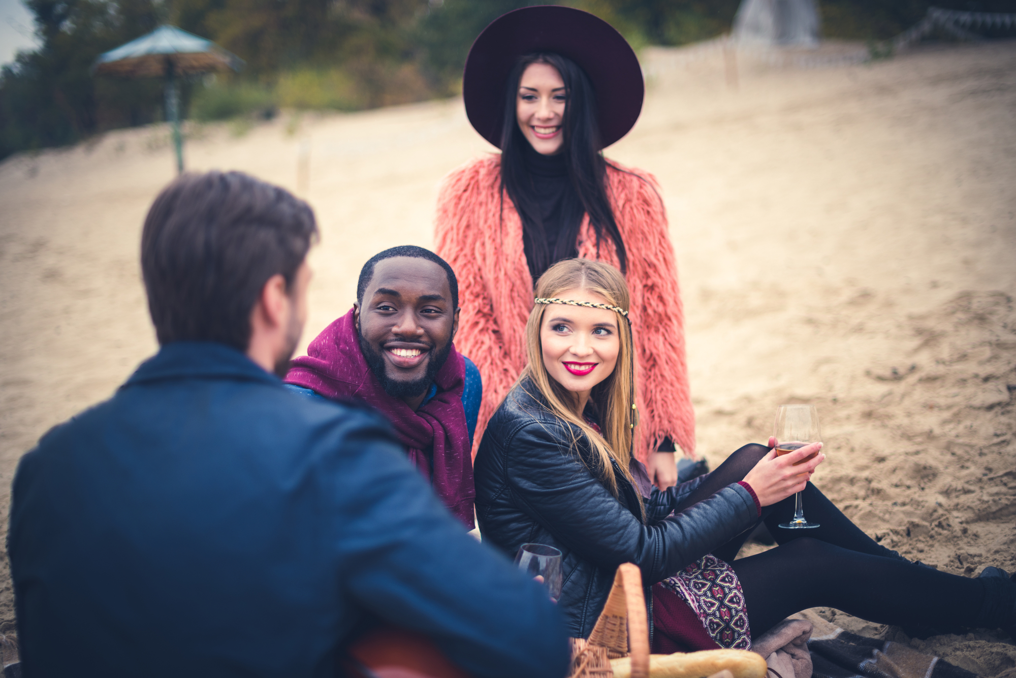 Four friends enjoying a day at the beach. Two are seated on the sand, one holding a wine glass, and the other two stand nearby. They are dressed in fall clothing, with one woman wearing a stylish hat and another a headband. They appear happy and engaged in conversation.