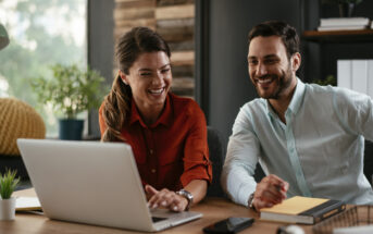 Two people are sitting at a desk and looking at a laptop screen while smiling. The person on the left is wearing a red shirt, and the person on the right is wearing a light blue shirt with a yellow notebook in front of them. They appear engaged and happy.