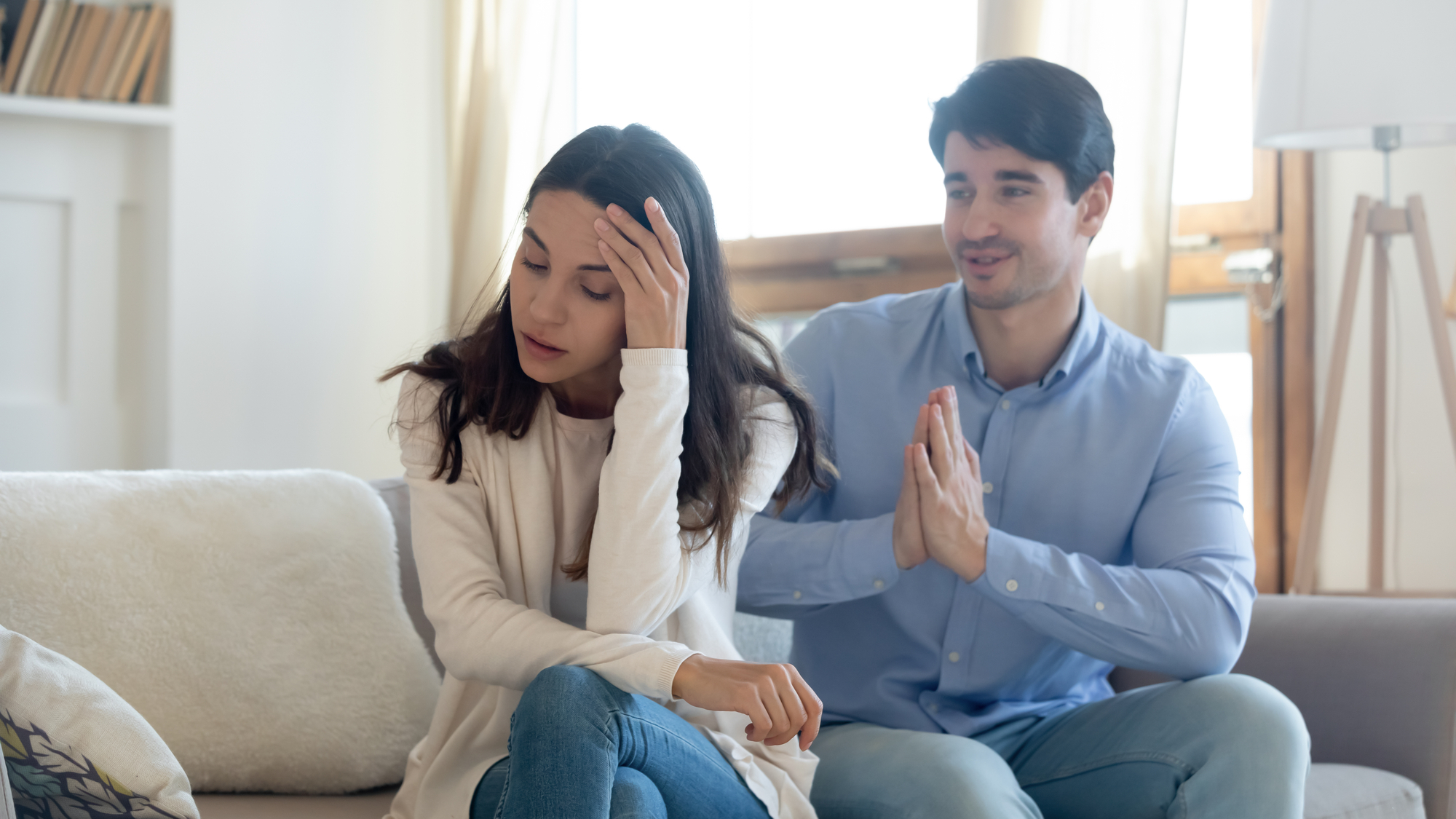 A woman sits on a couch with her hand on her forehead, appearing upset. A man next to her clasps his hands together, as if pleading or apologizing. Both individuals are casually dressed, and the room appears bright with large windows in the background.