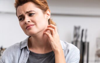 A woman with a concerned expression scratches her neck with her left hand. She has light brown hair pulled back and is wearing a grey shirt with a lighter grey cardigan. In the blurry background, there is a set of kitchen knives.