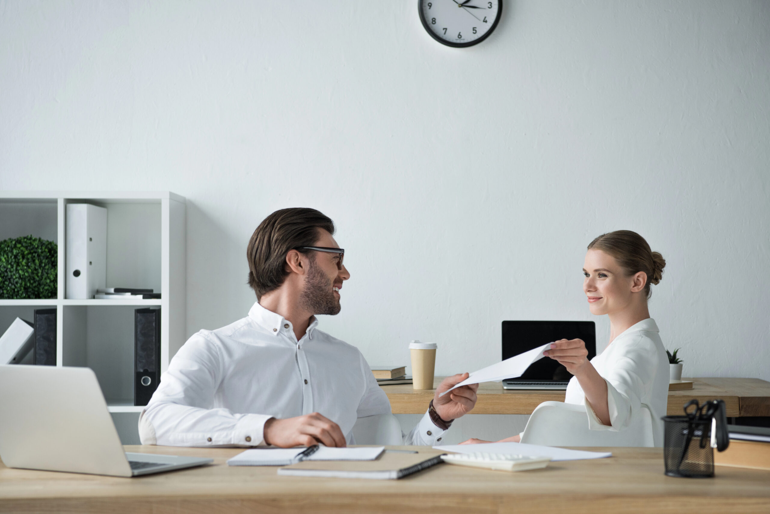 In a modern office, a smiling man in glasses and a white shirt sits at a desk, passing a sheet of paper to a woman with blonde hair and a white blazer sitting opposite. Office decor includes a clock, shelving, and laptops.