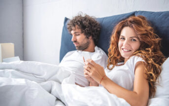 A woman with long curly red hair smiles at the camera while holding a phone, lying in bed with white sheets. Next to her, a man with curly dark hair focuses on his own phone. They are both dressed in white tops, and the background is a white wall.